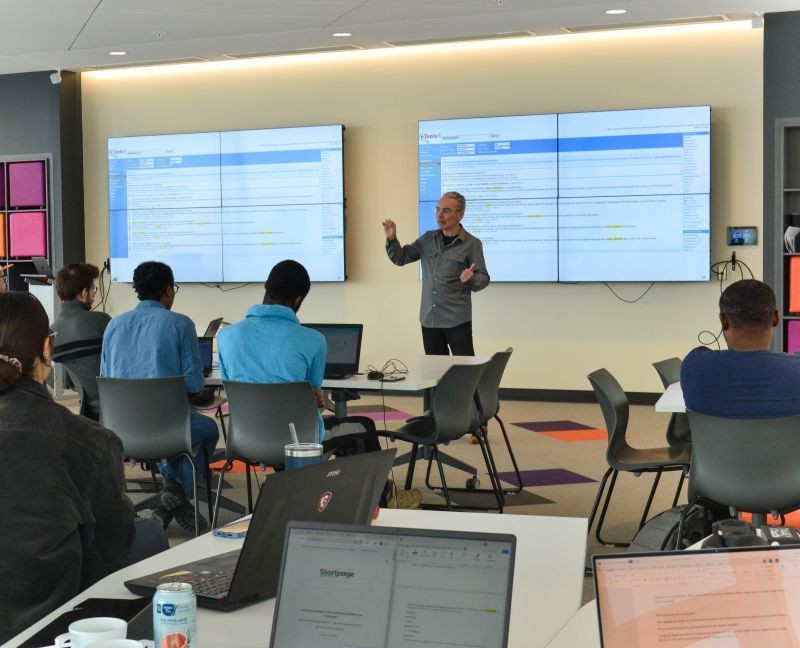 Person giving a presentation in a classroom with large screens and attendees using laptops.